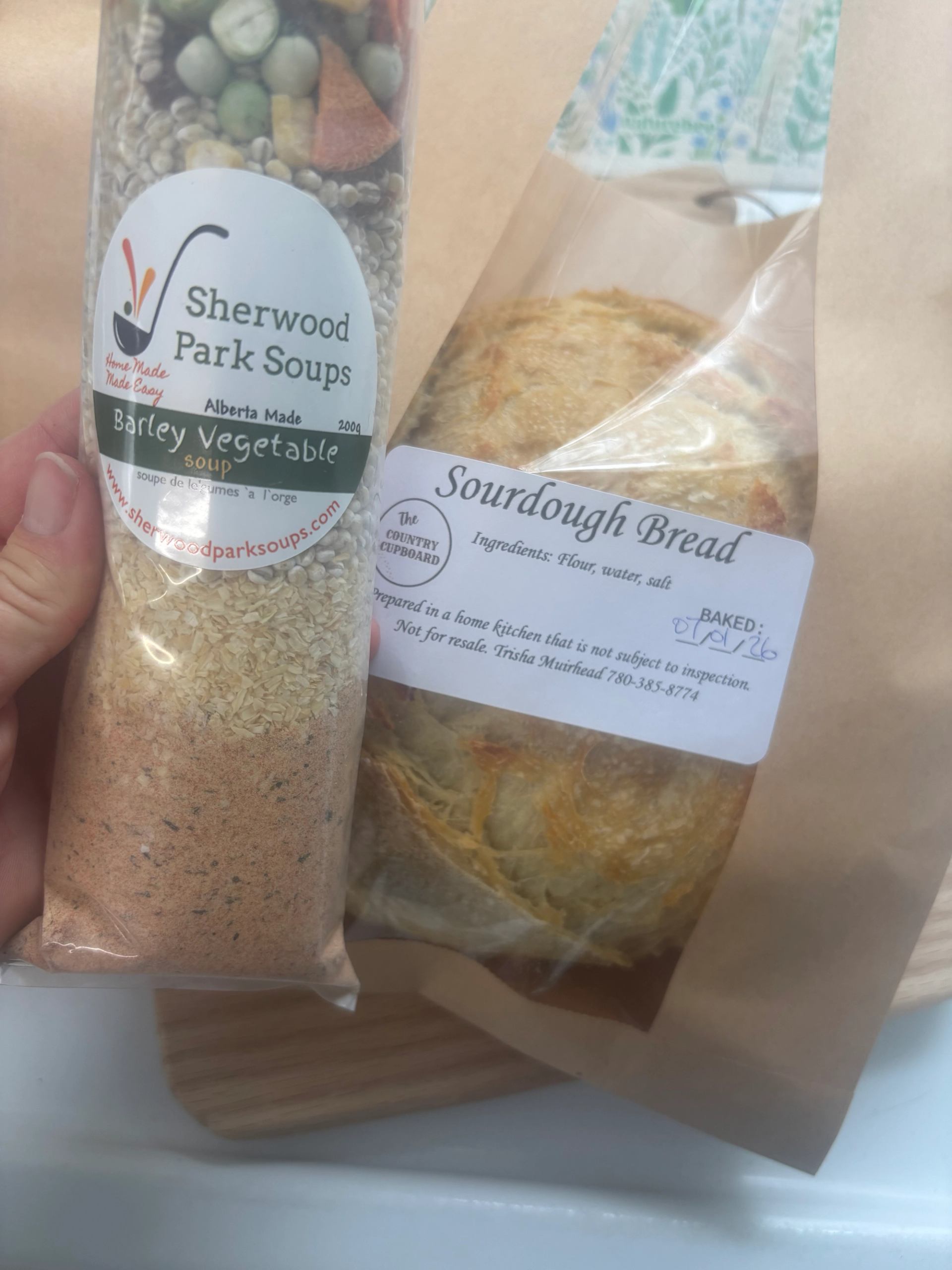 A hand holds a bag of Sherwood Park Barley Vegetable soup mix next to a sourdough bread loaf in a brown paper bag.