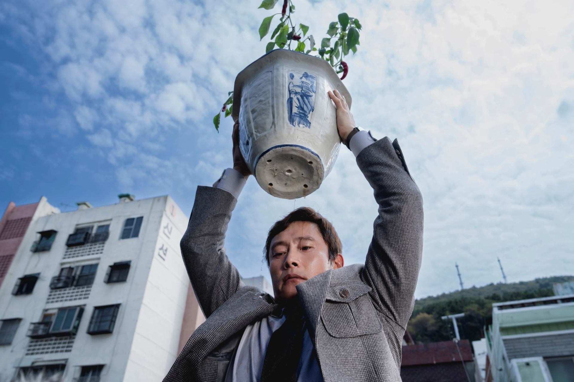 A person holds a large potted plant overhead outdoors, with apartment buildings and sky in the background.