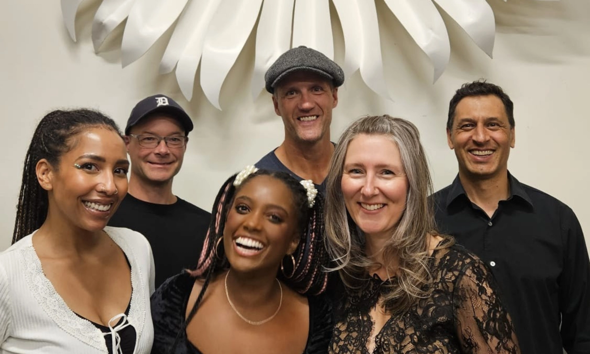 Group of six adults posing indoors in front of a large white wall sculpture.