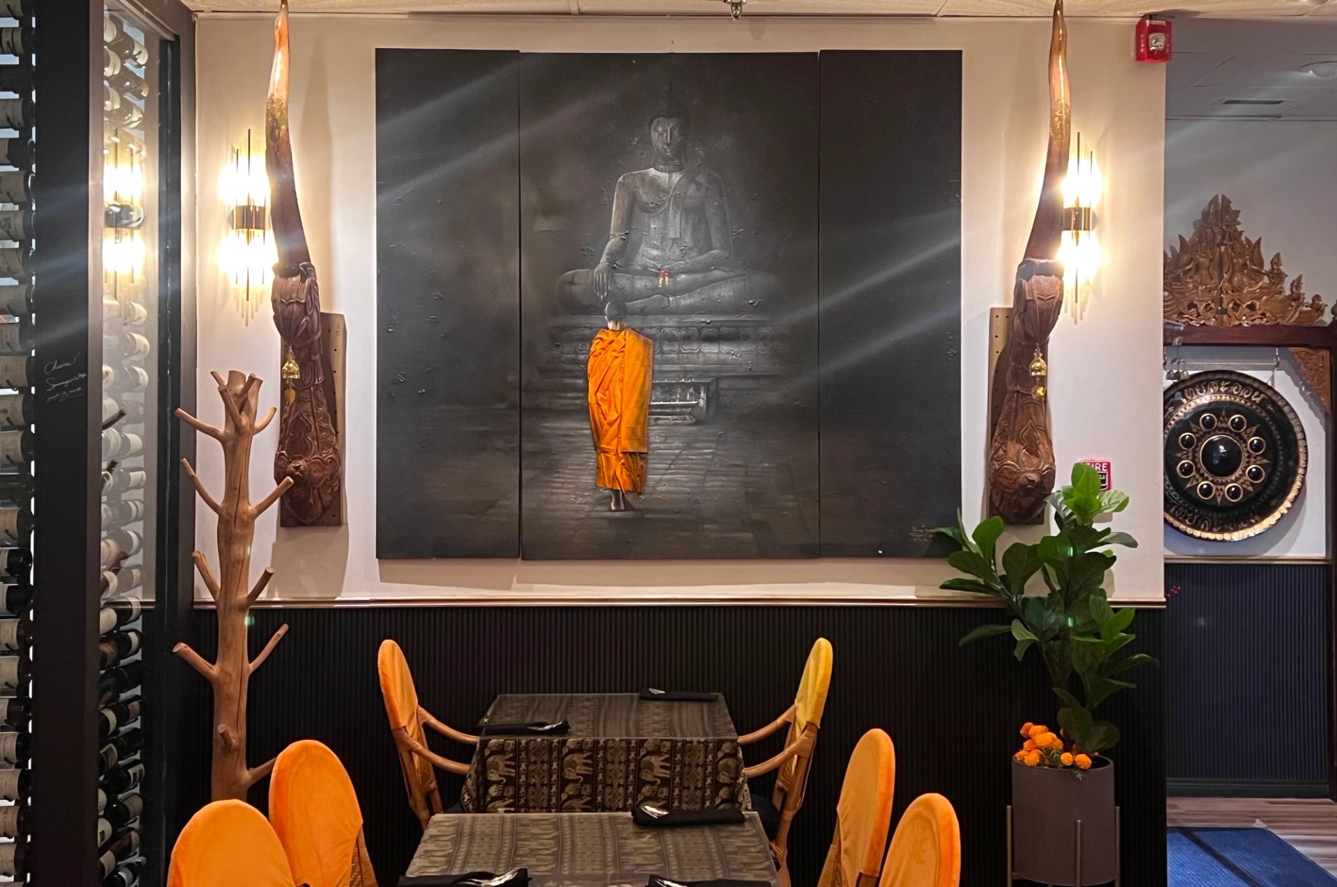 Restaurant dining area with Buddha artwork, orange chairs, carved wood and decorative gong.