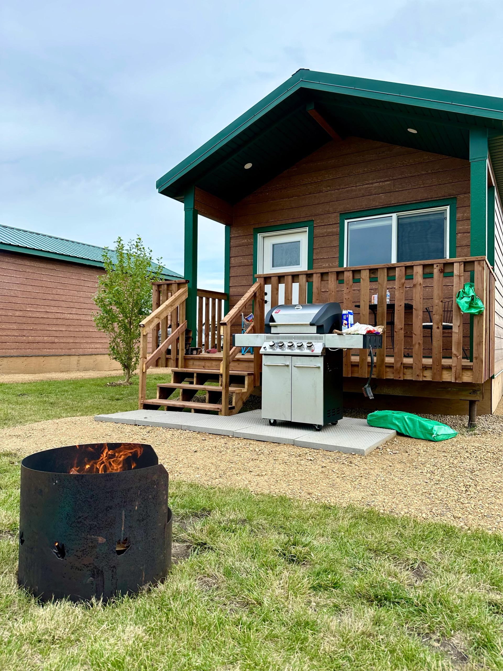 Cabin with porch grill, fire pit, and green roof in grassy campground setting.