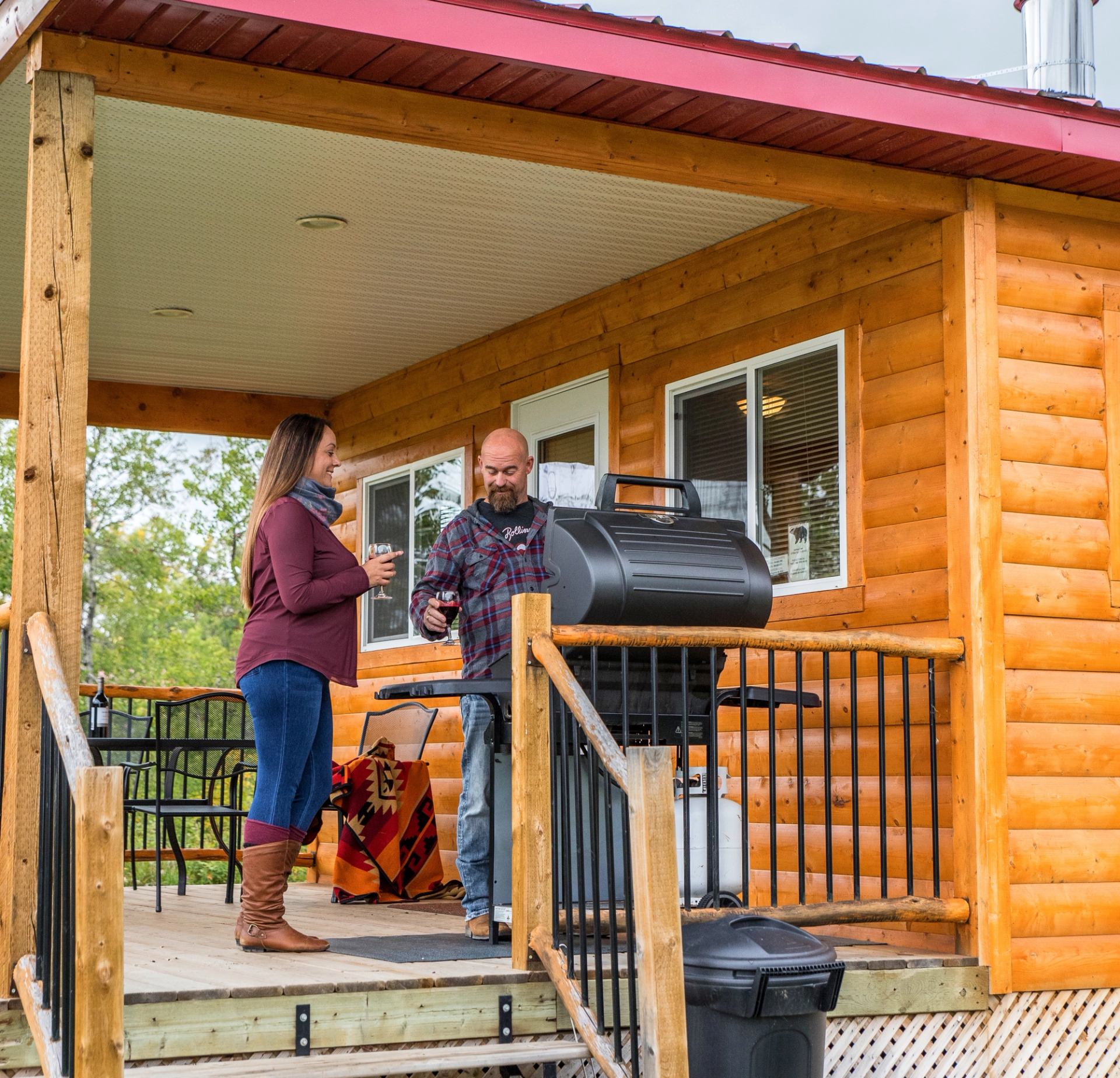 Two people standing on a wooden porch grilling near a log cabin with railing