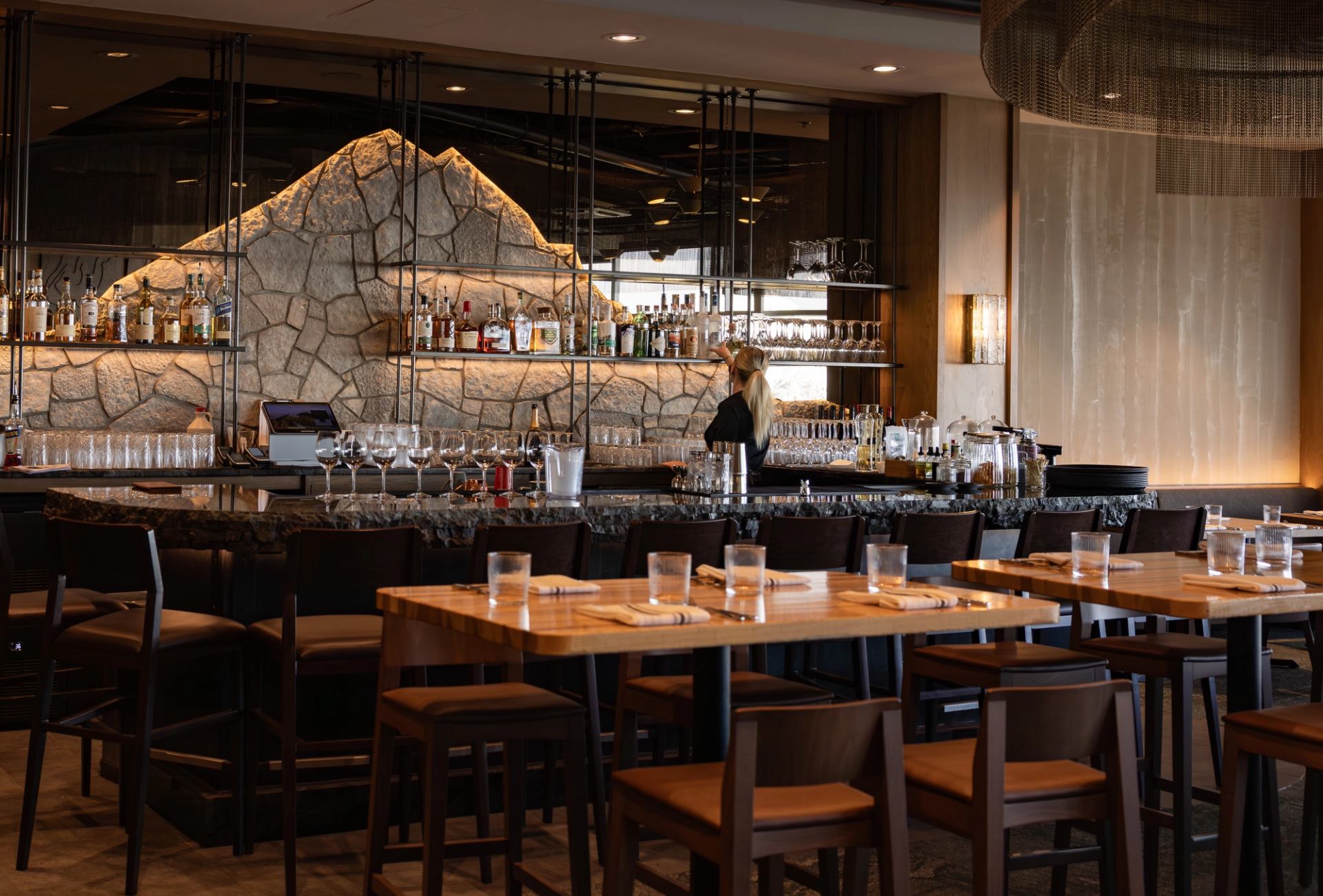 Restaurant bar with stone feature wall, bartender arranging glassware, and set wooden tables.