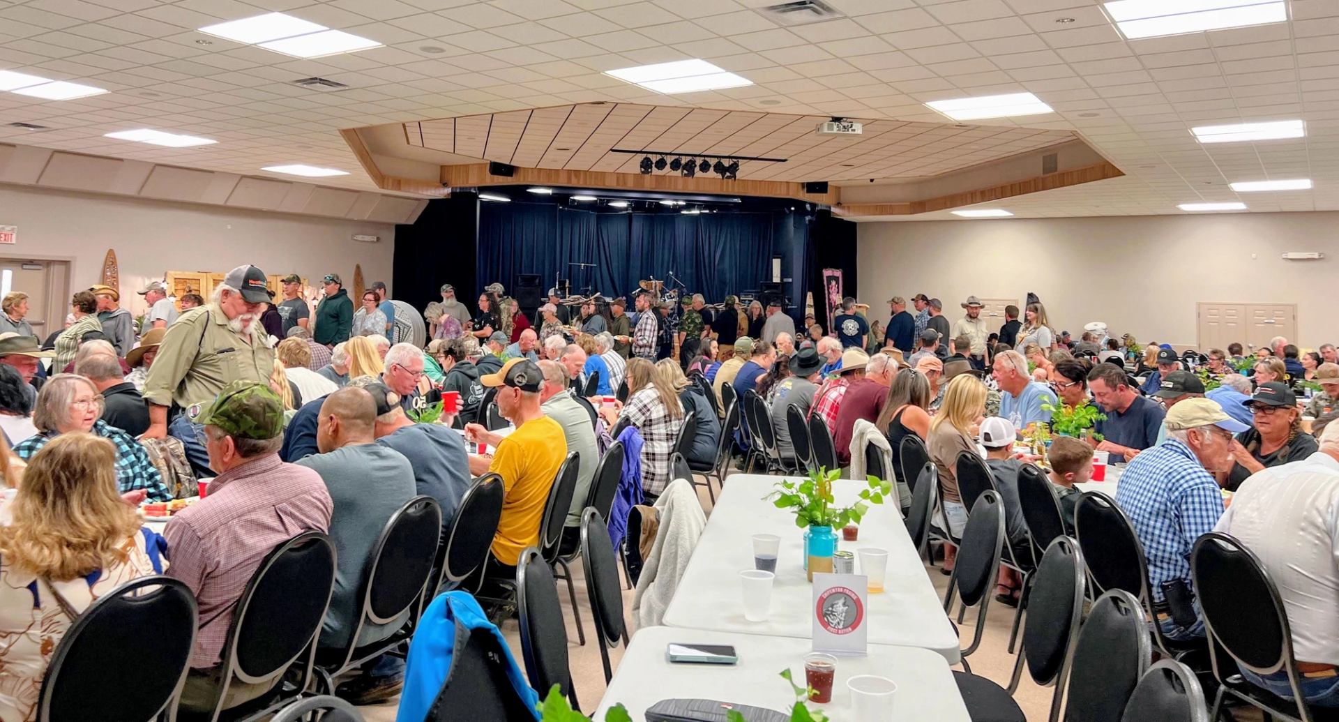 Crowded community hall with people seated at long tables eating and talking together.