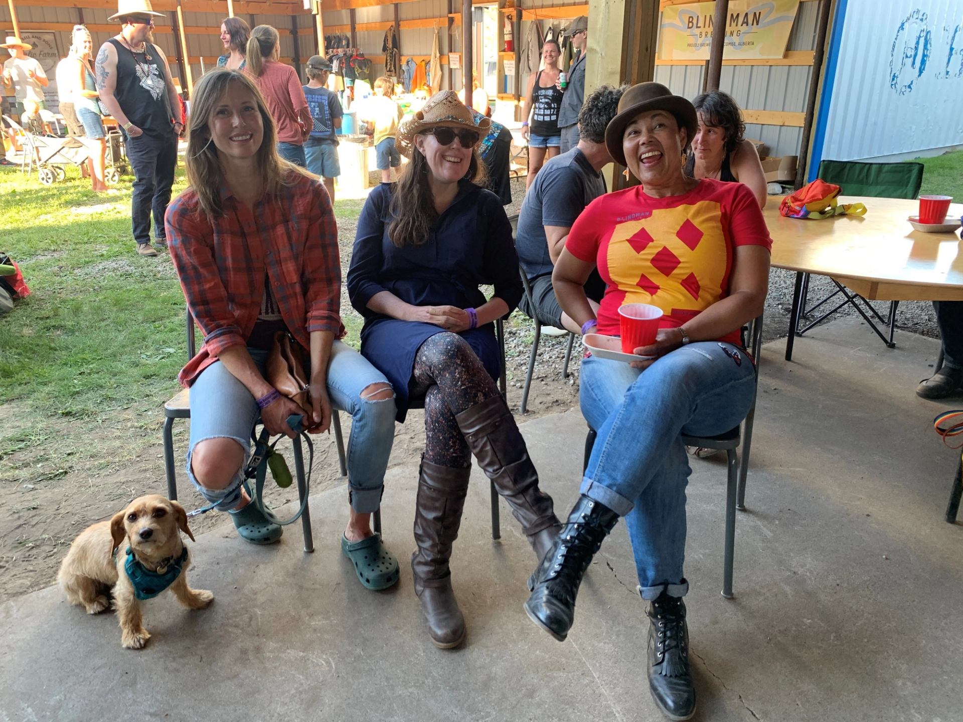 Three smiling women sit on chairs with a small dog by their feet in a casual setting.