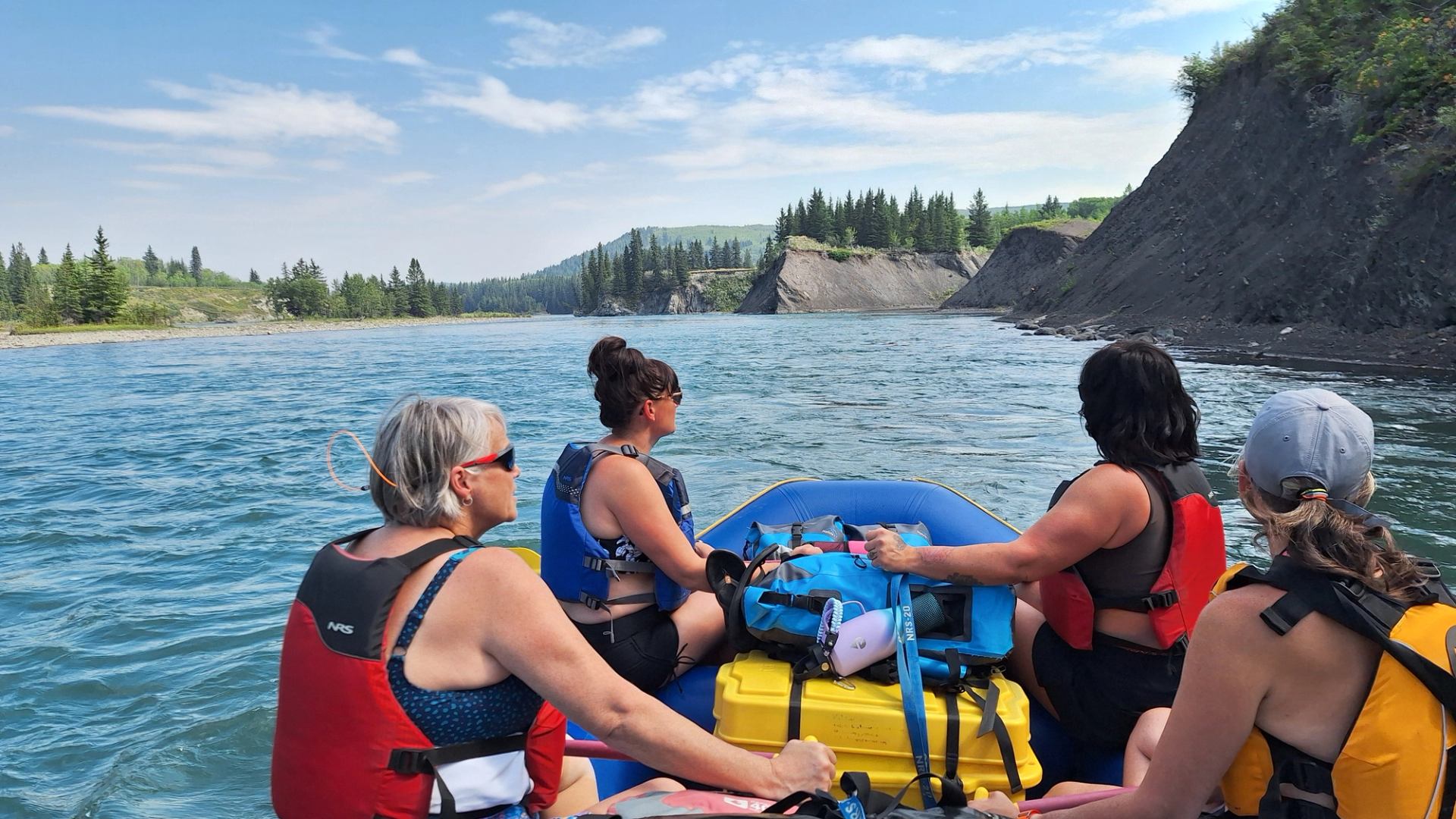 Group rafting on a wide river with forested cliffs under a bright blue sky.