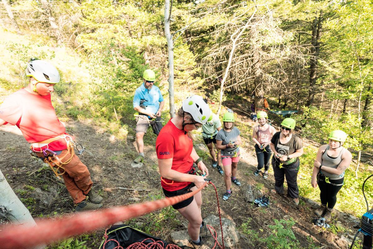 Rock climbing group wearing helmets and harnesses gathers on forested slope with ropes.