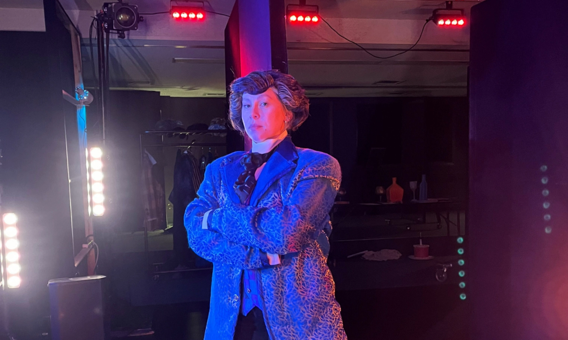 Performer in patterned blue suit stands under stage lights with arms crossed backstage.