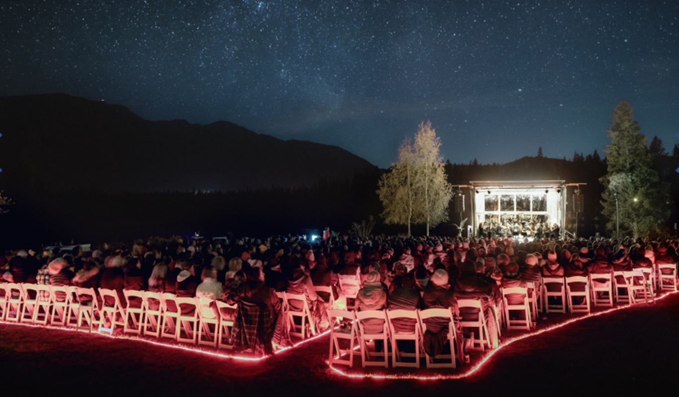 Outdoor audience watches illuminated stage concert under starry night sky and mountain silhouette.