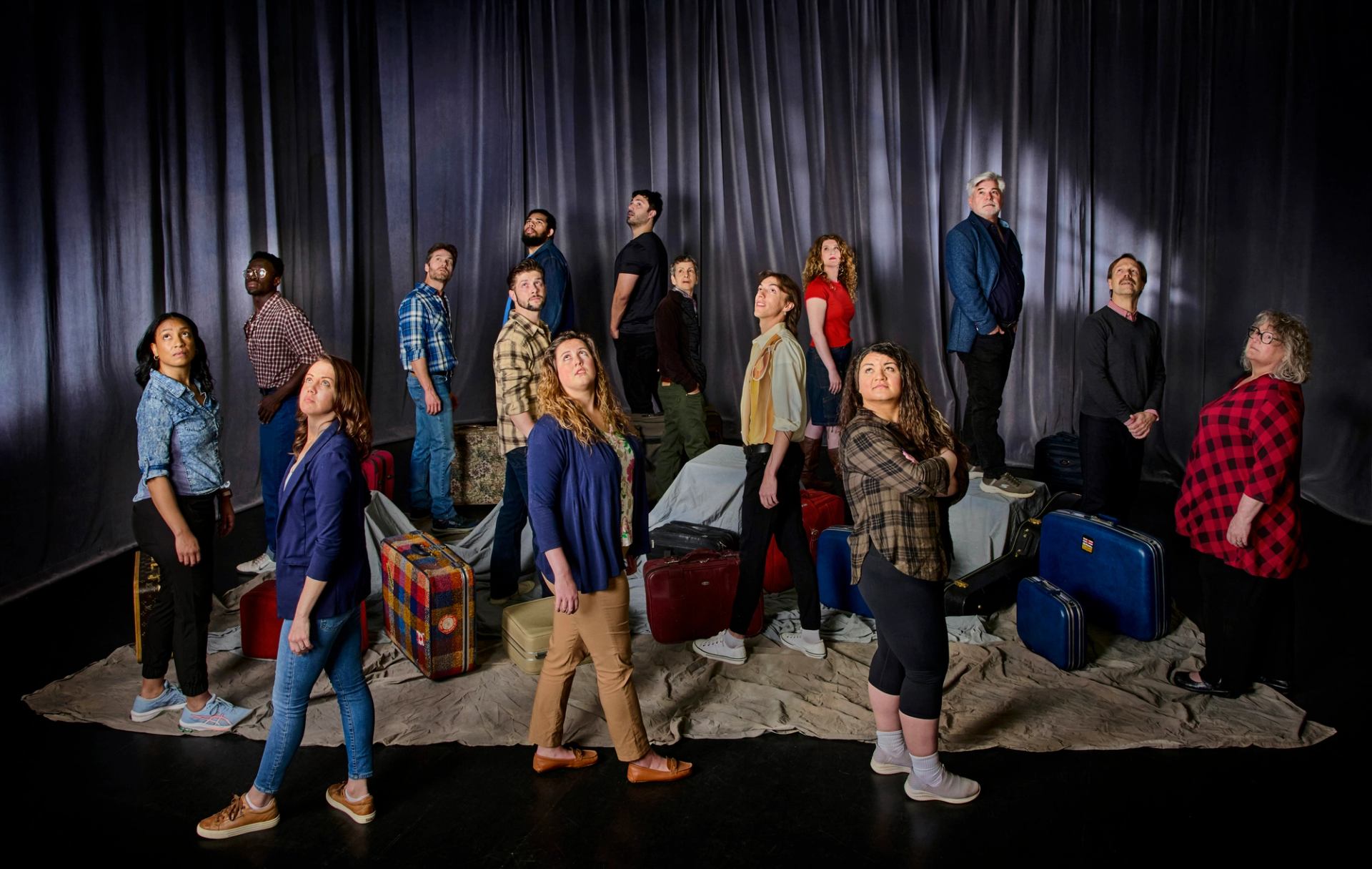 Group of people stand among stacked suitcases onstage, looking upward before grey curtains.