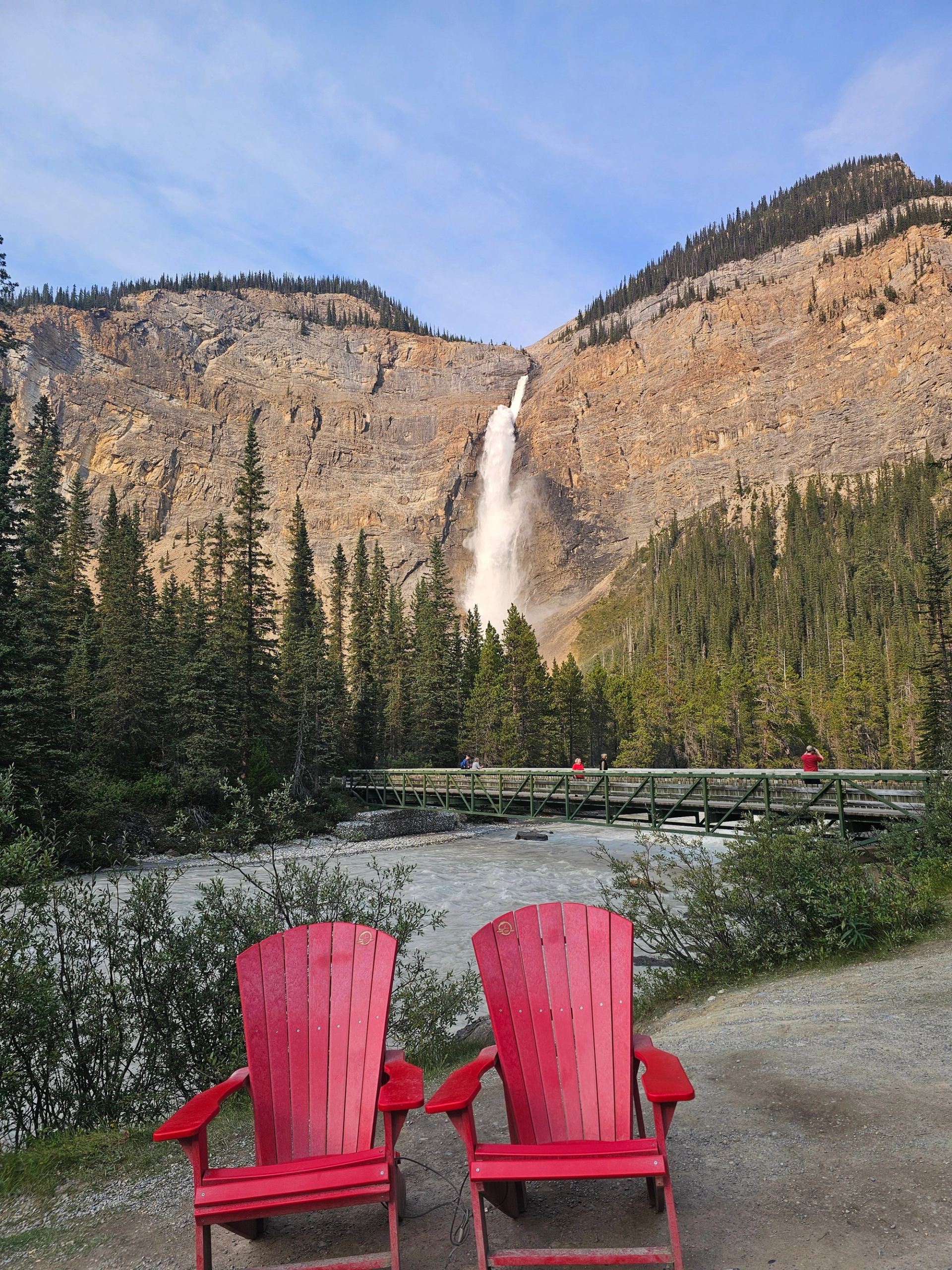 Red Adirondack chairs facing tall waterfall, forested mountains and footbridge over river.