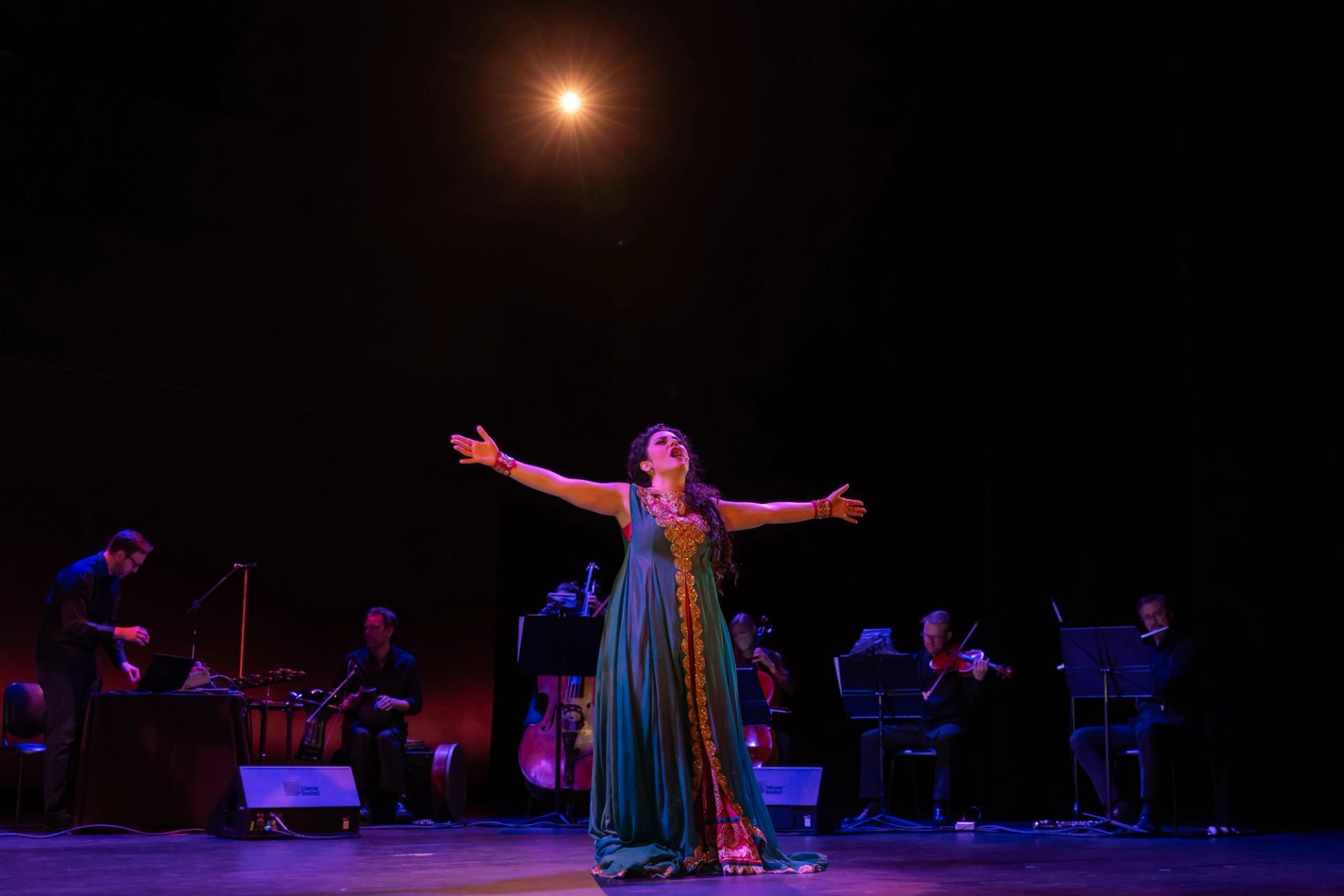 Opera singer in spotlight with musicians on dimly lit stage.