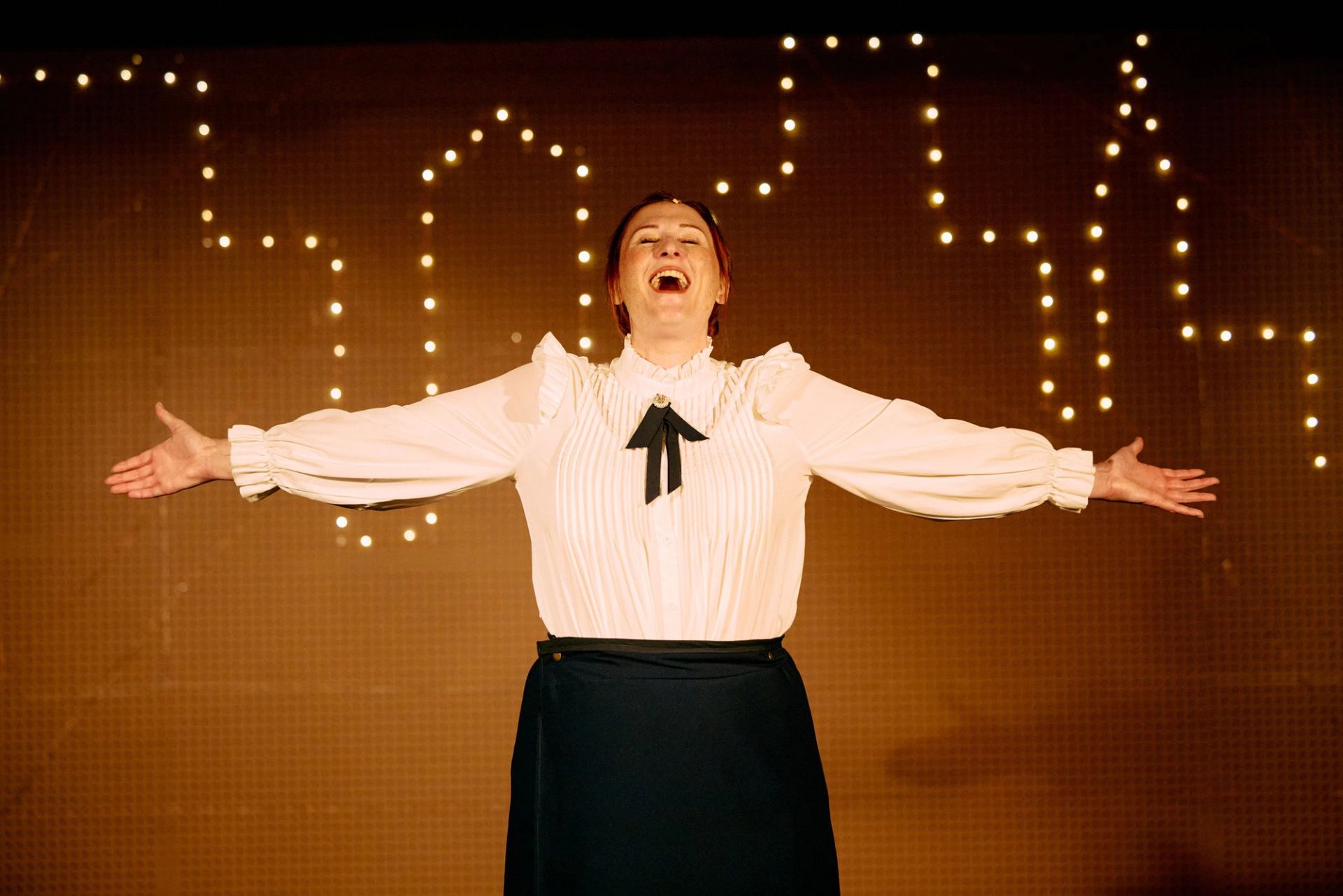 Performer in white blouse stands on stage, arms outstretched, with string lights behind.