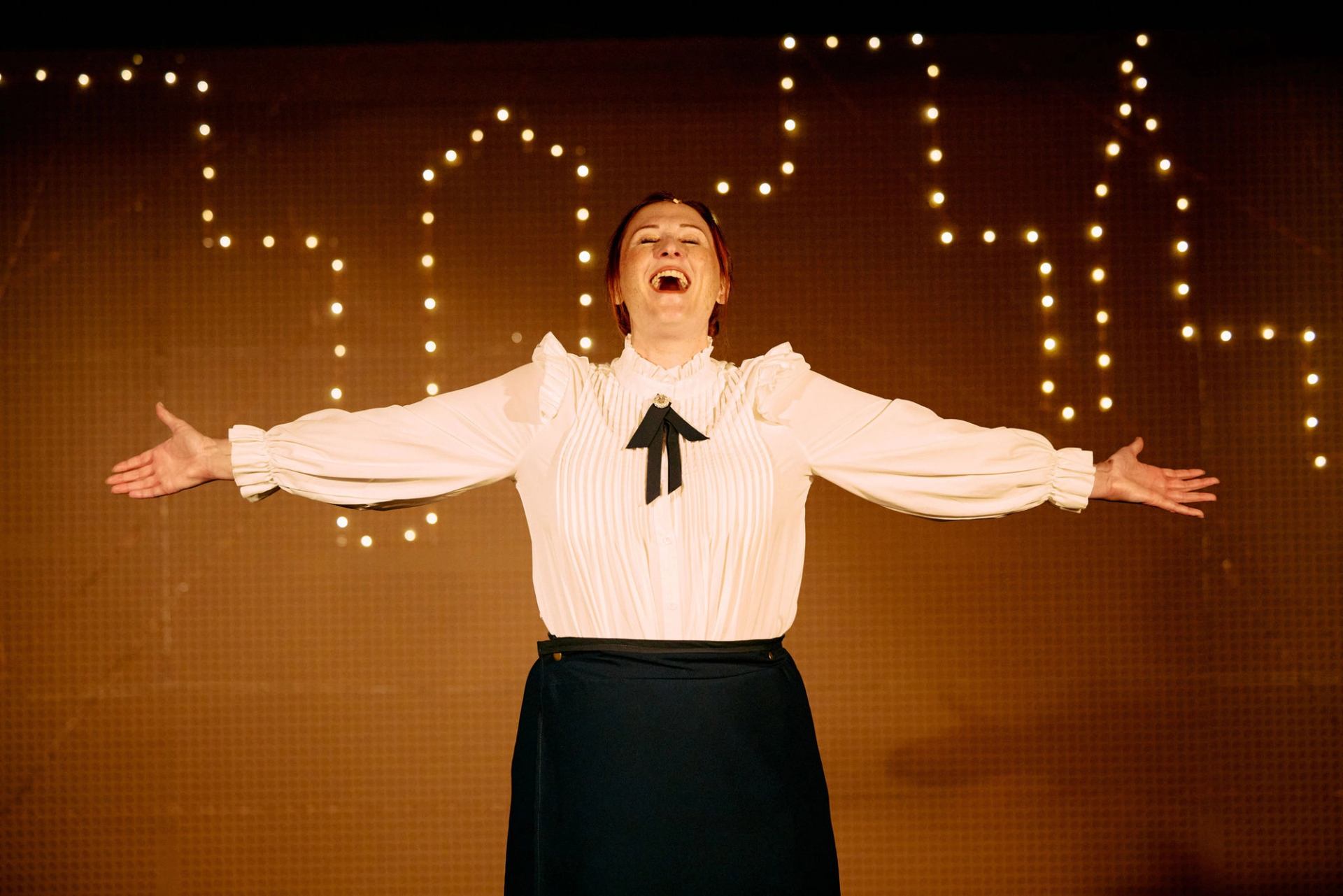 Performer in white blouse stands on stage, arms outstretched, with string lights behind.