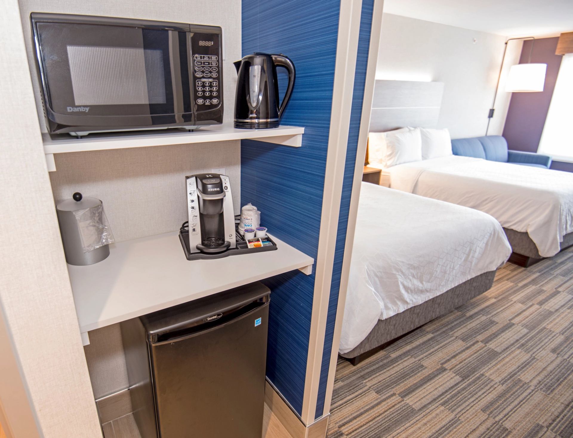 Modern hotel room with a kitchenette on the left, featuring a microwave, kettle, coffee maker, and mini-fridge. To the right, a cozy bed with white linens. Neutral tones create a welcoming ambiance.