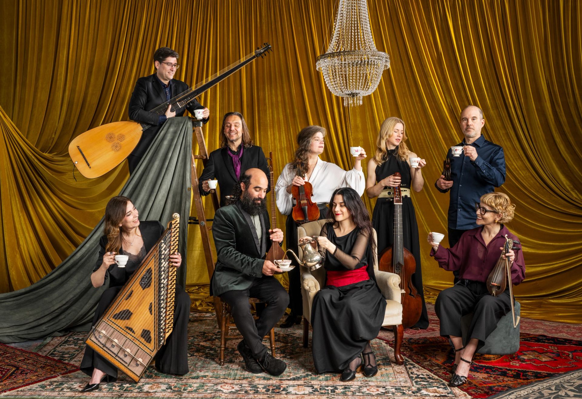 Musicians with string instruments share tea under a chandelier against gold curtains.