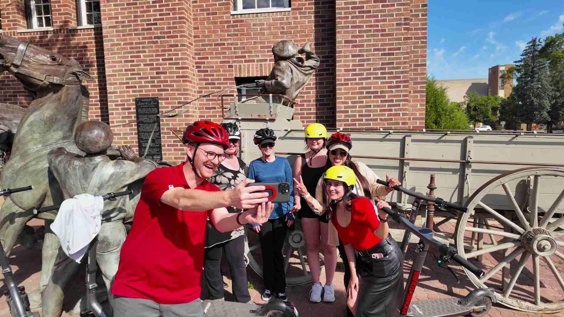 Group of scooter riders in helmets taking a selfie beside brick building and statues.