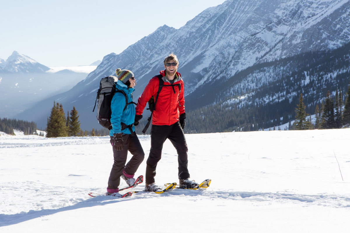 Two people snowshoe across open snowfield with backpacks, mountains and trees in background.