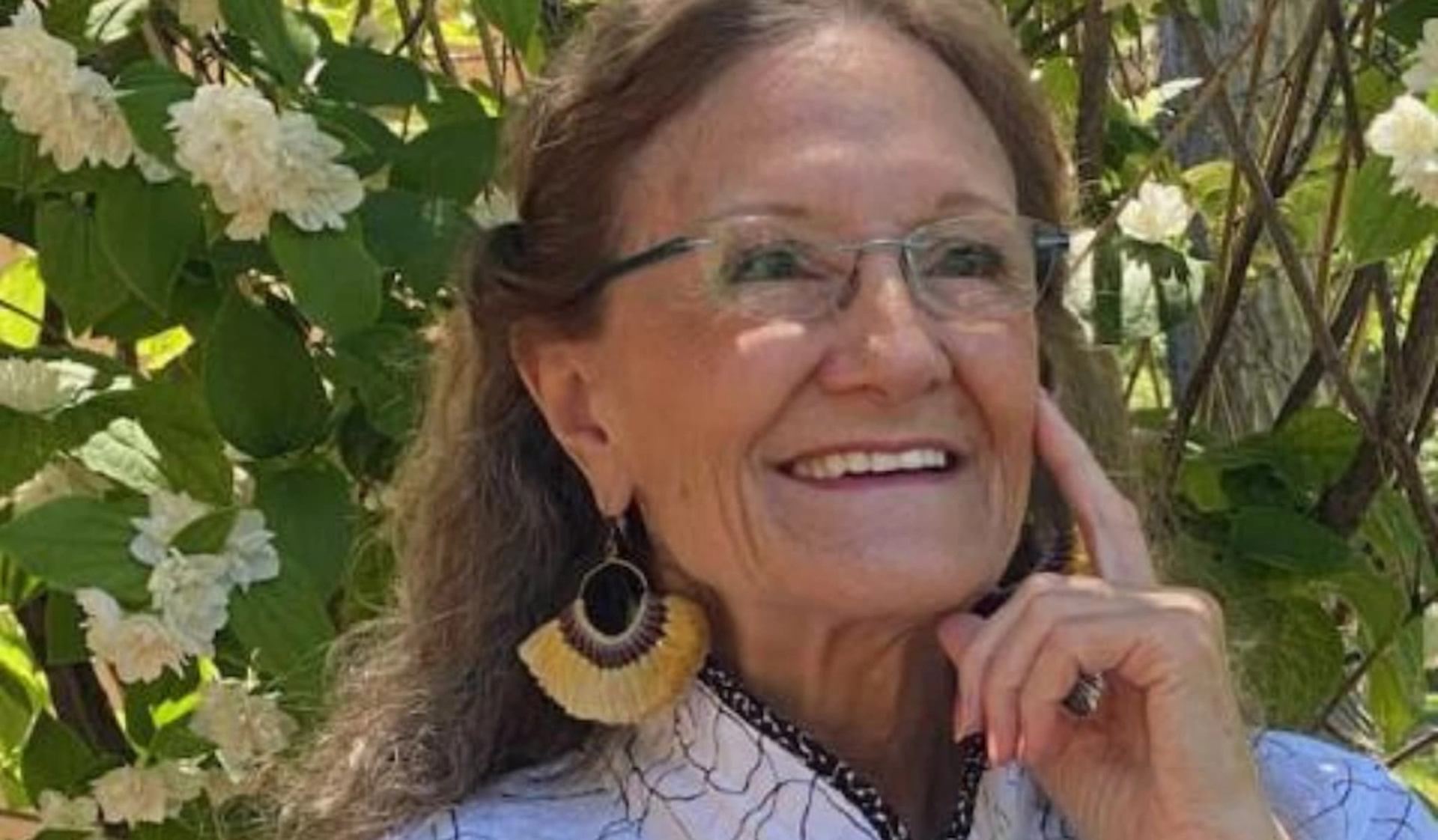Person wearing glasses and large earrings stands near leafy bushes with white flowers.