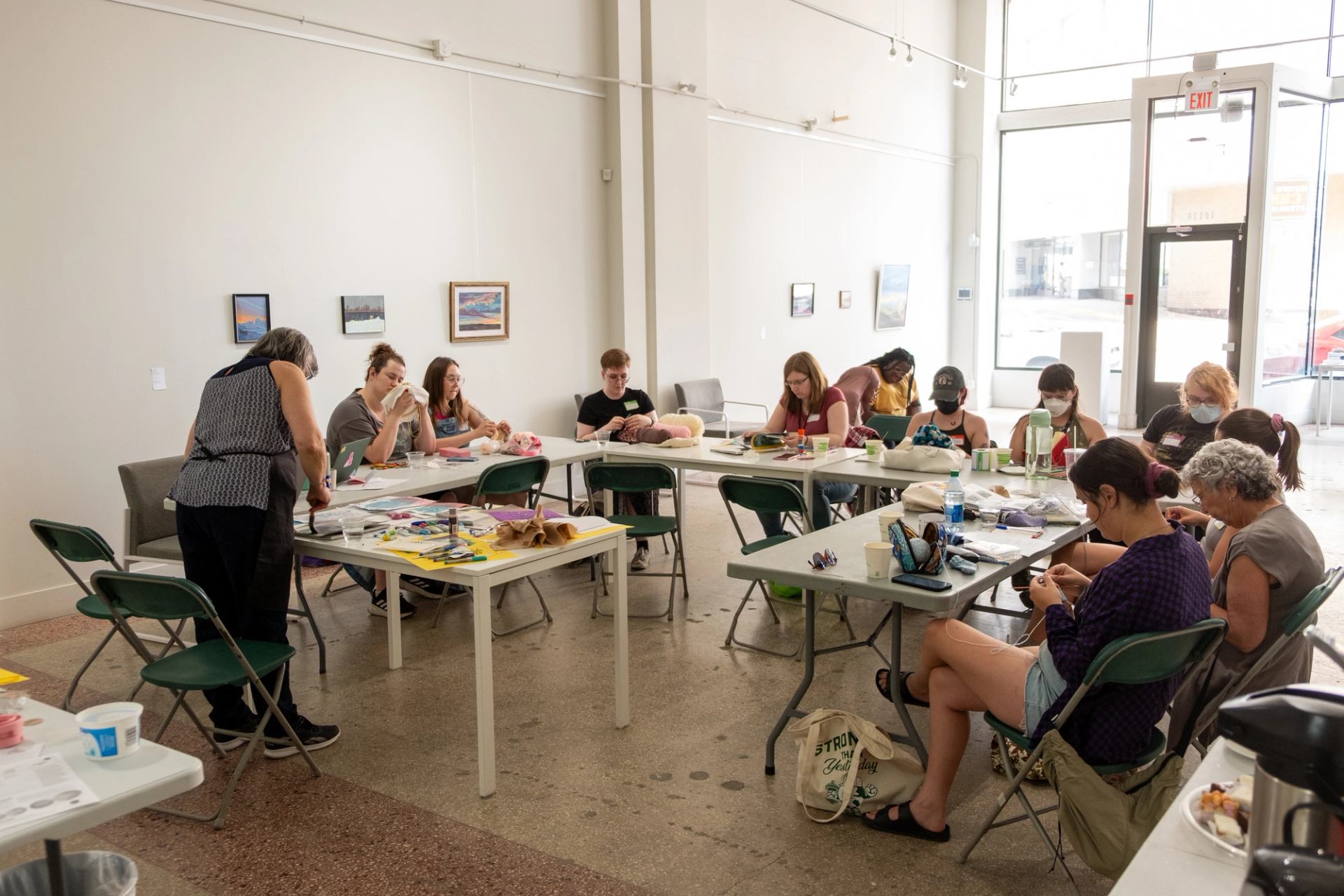 In the gallery around folding tables, people sit working on crafts. The gallery is brightly lit.
