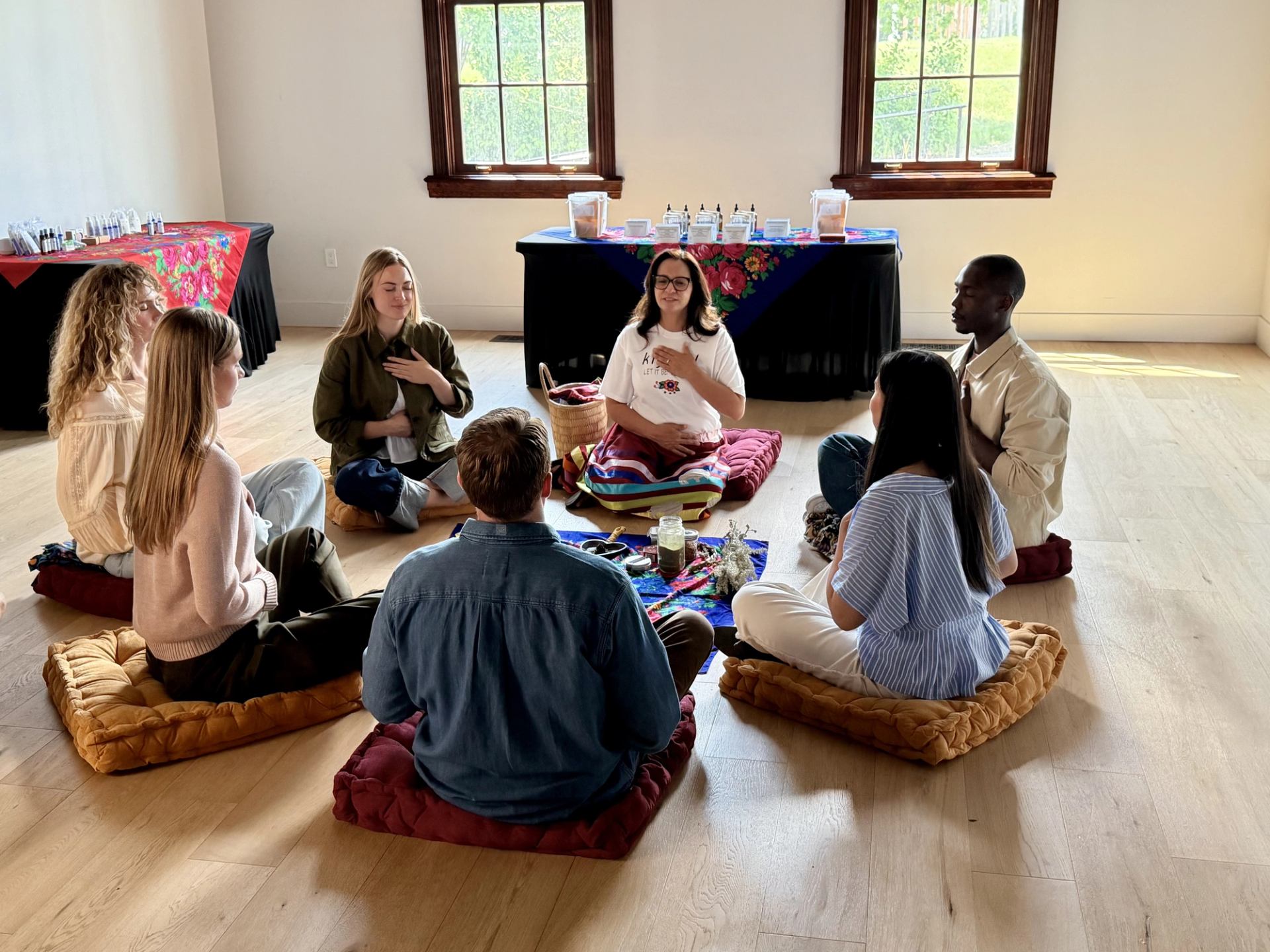 Group of people sit on floor cushions in a circle, meditating together indoors.