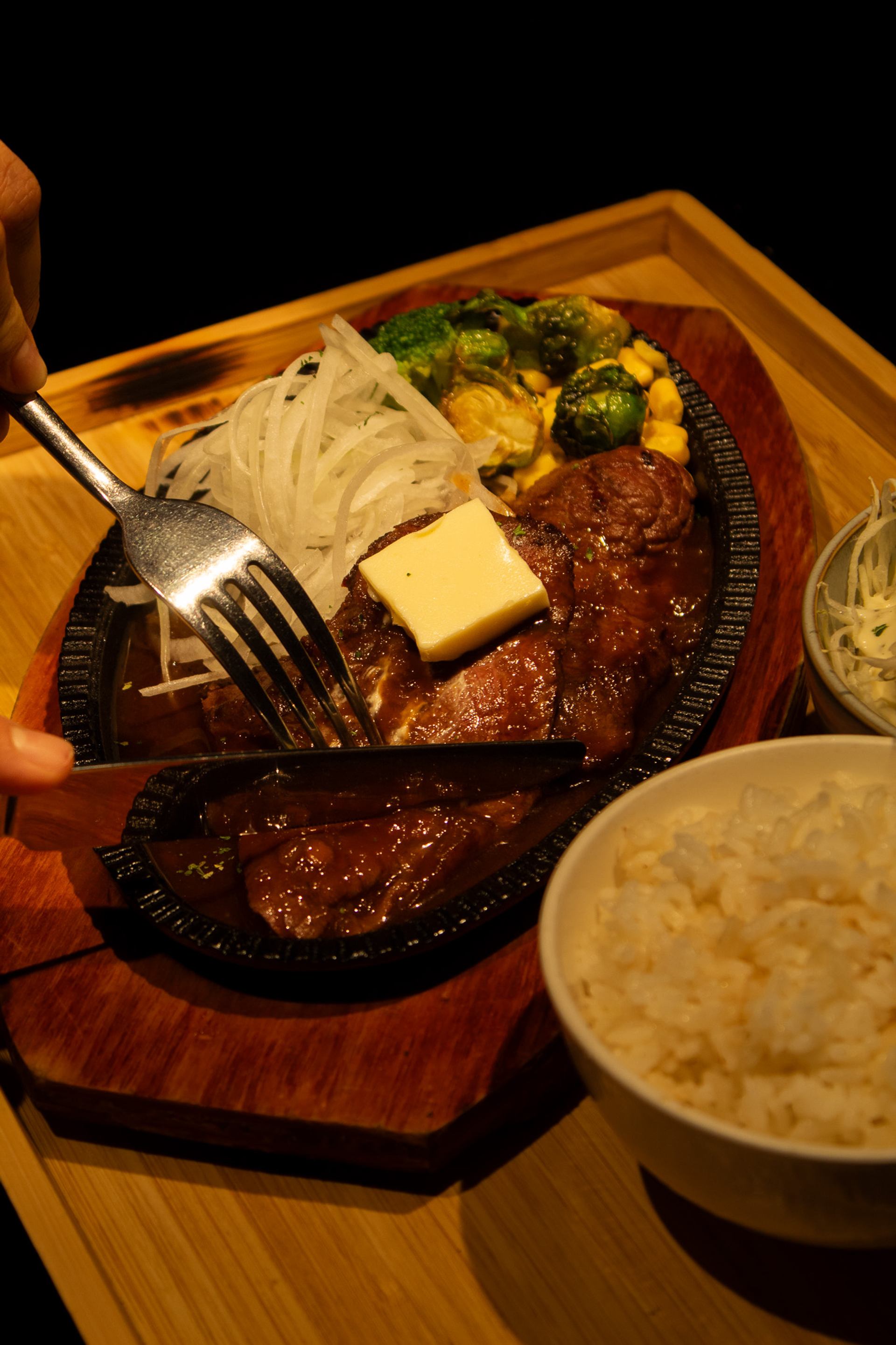 Sizzling steak with butter, onions and vegetables on hot plate beside bowl of rice.