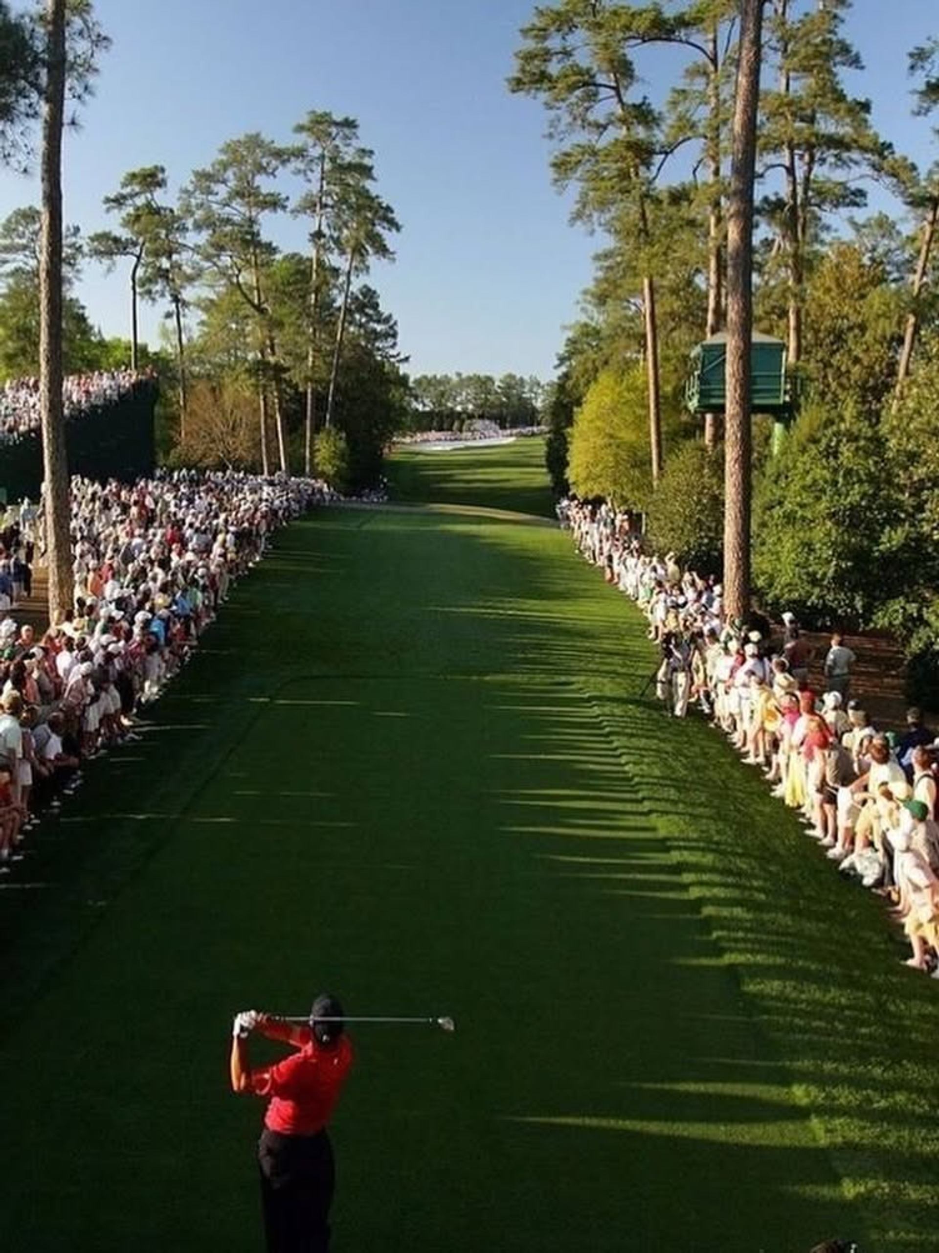 A golfer tees off along a tree-lined fairway with spectators gathered on both sides of the course.