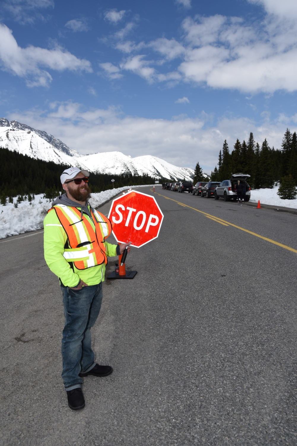 Volunteer in safety vest holds red STOP sign by highway with mountains and vehicles.