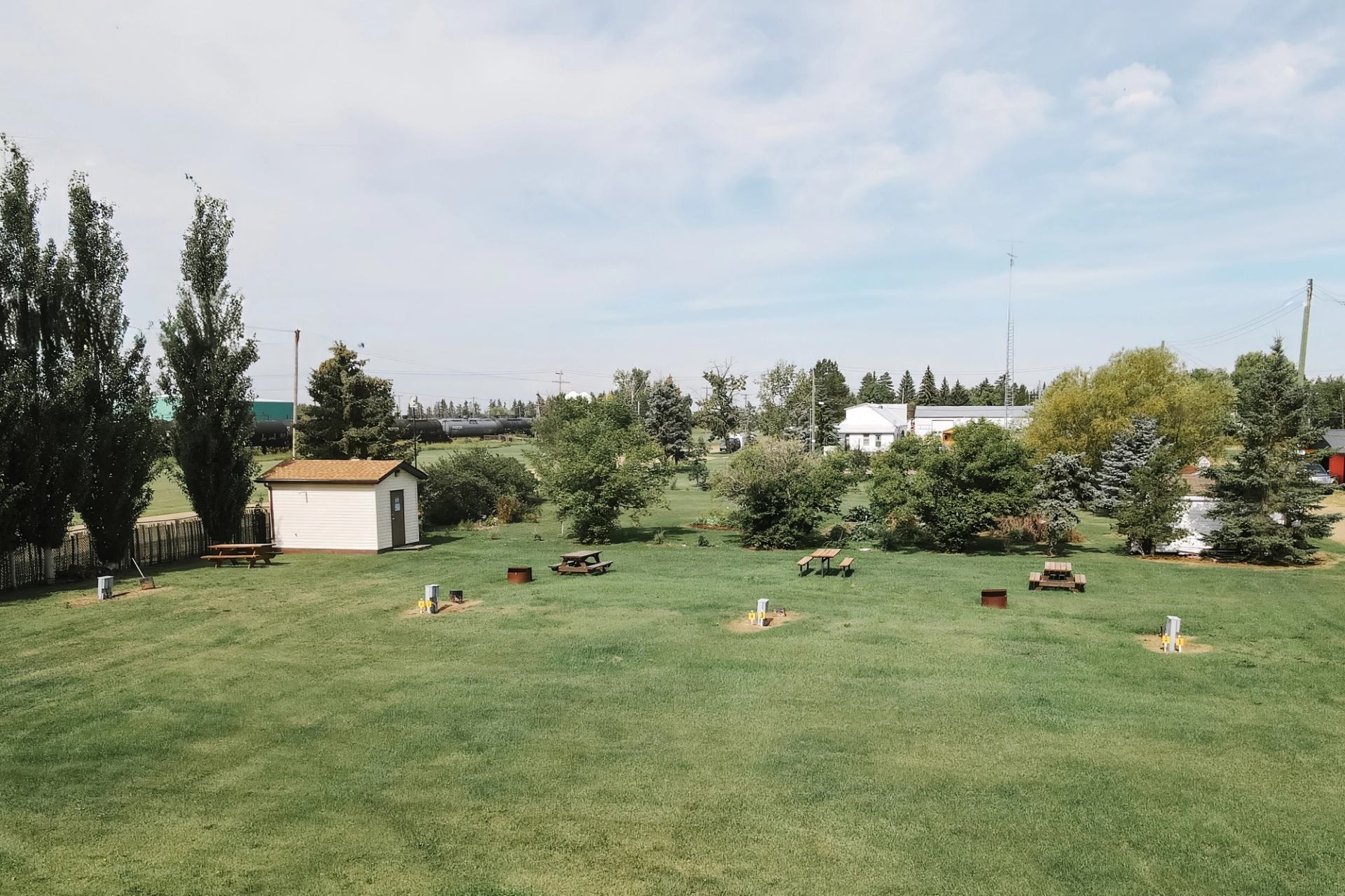 Grassy campground with picnic tables, fire pits, small shed and surrounding trees.