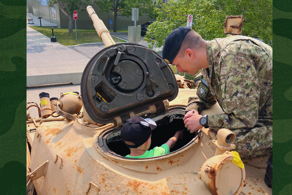 Person in camouflage repairs equipment inside an open military vehicle hatch outdoors.