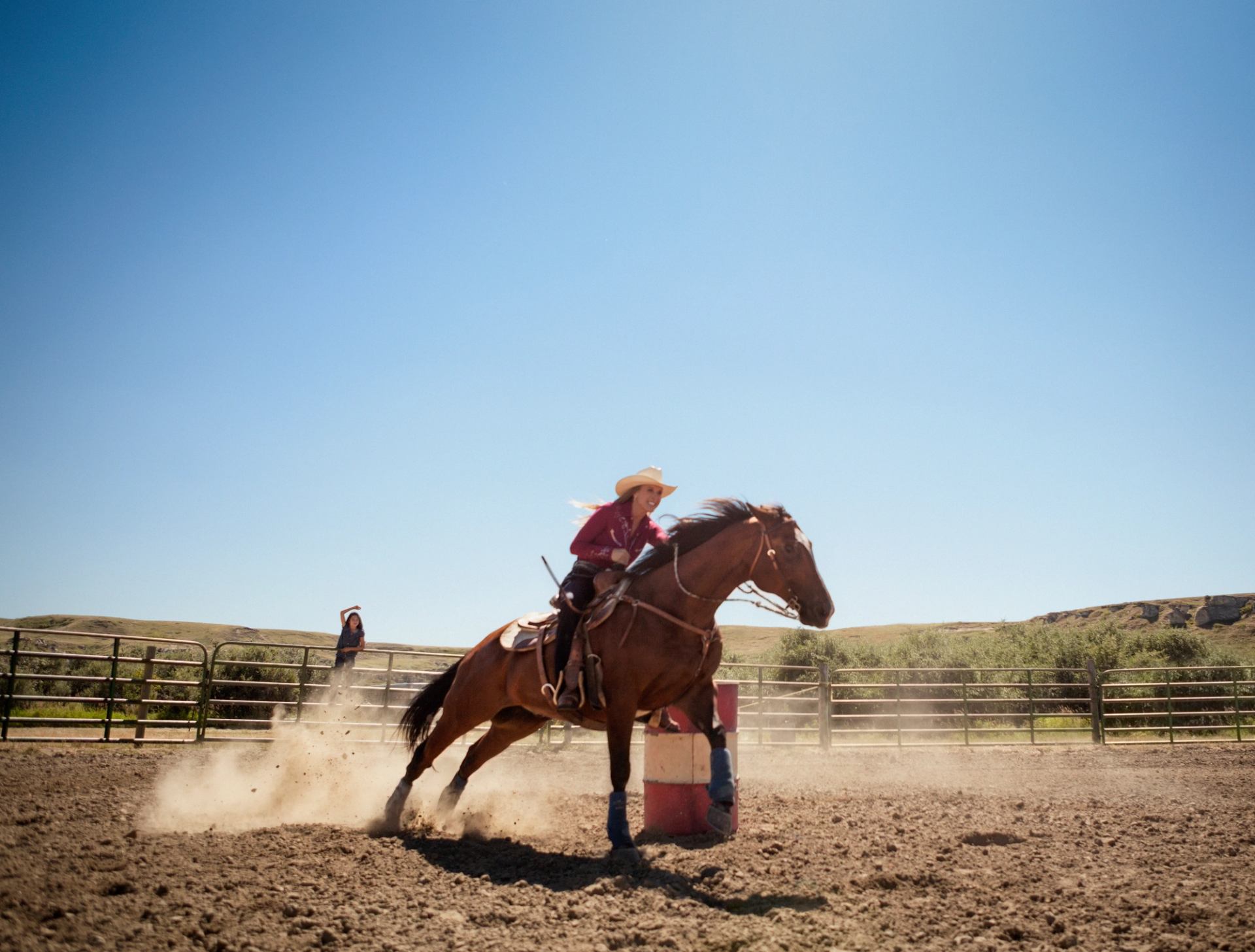 Rider on galloping horse rounds a barrel in dusty outdoor arena under clear sky.