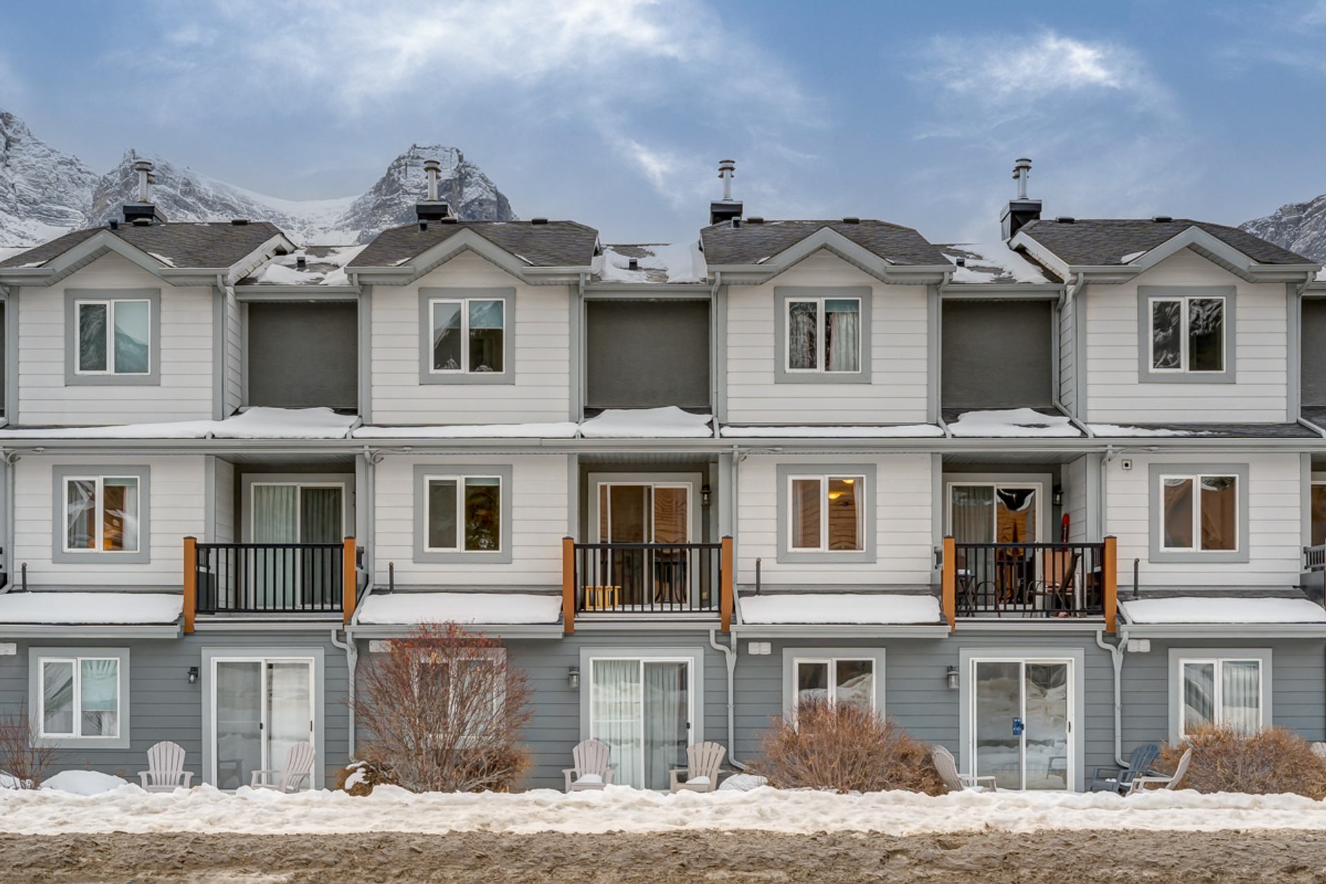 Snow-covered townhouse row with balconies, patio chairs and mountain peaks in background.