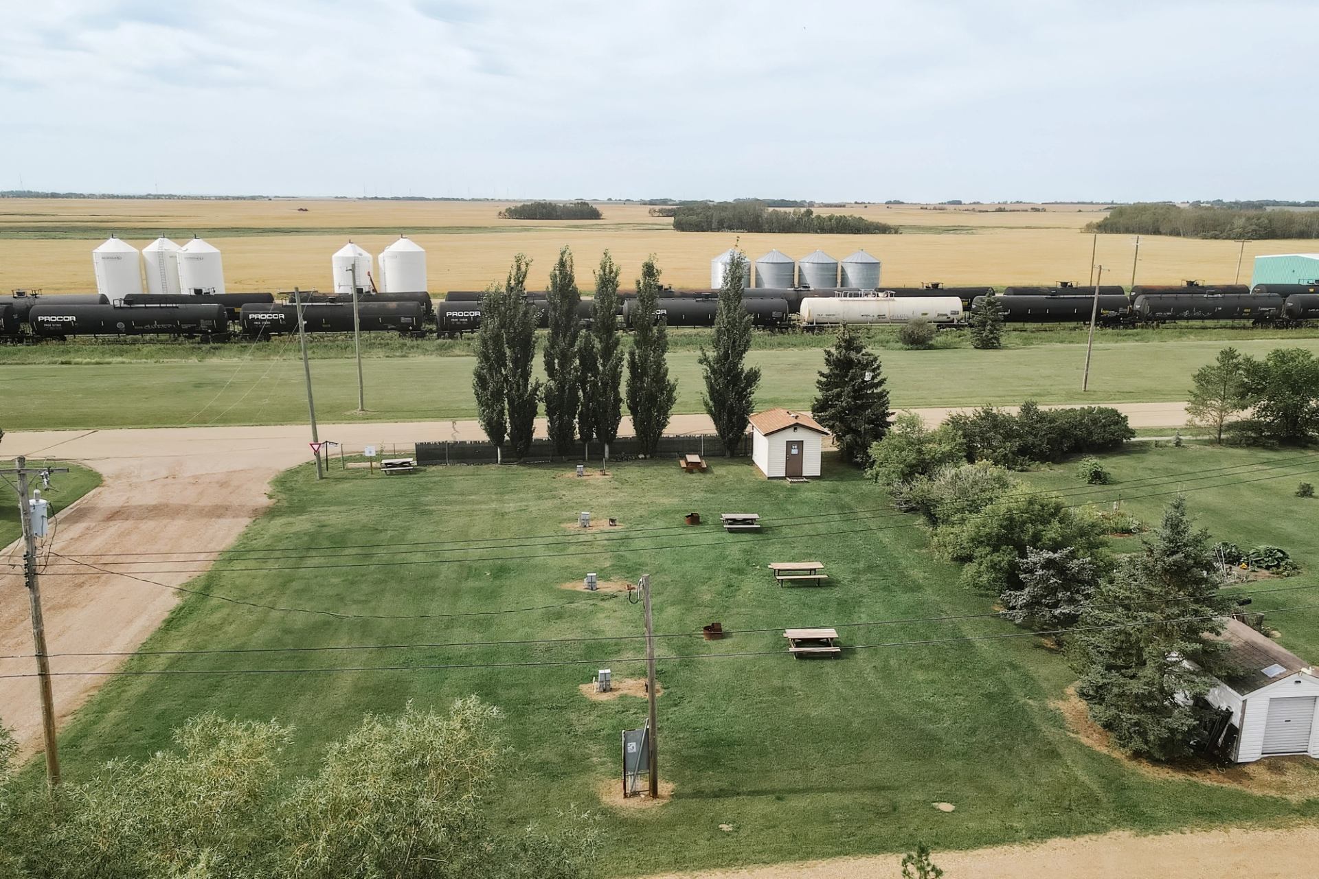 Railway tank cars pass grain silos behind grassy campground with picnic tables and trees.