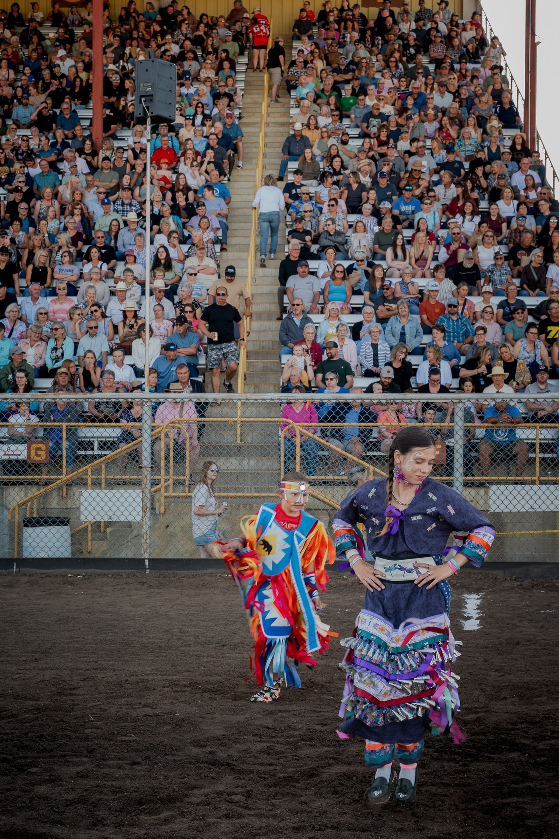 Powwow dancers in colourful regalia perform on dirt arena before large seated crowd.