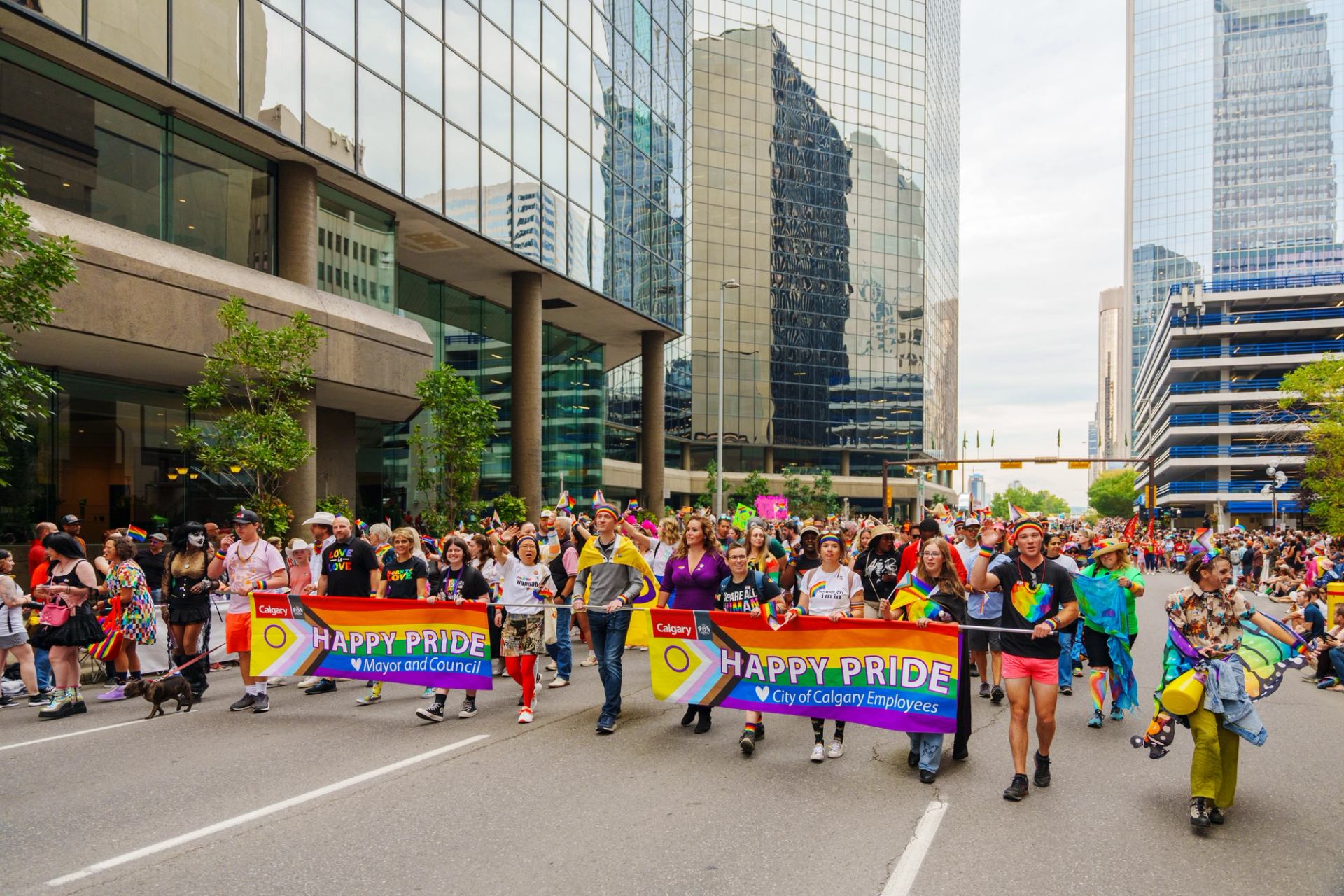 Large group holding Happy Pride banners between tall Calgary buildings.