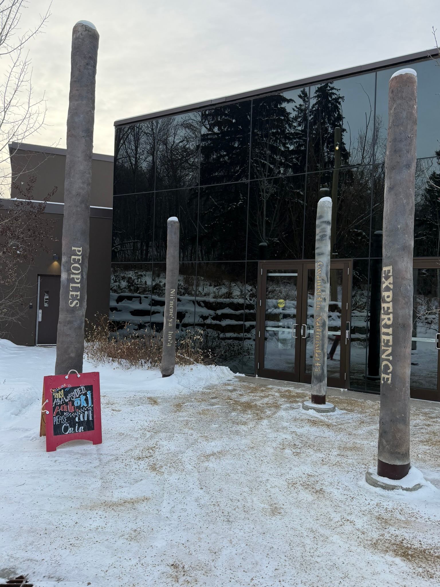 Snowy building entrance with tall sculpted pillars and reflective glass wall showing trees.