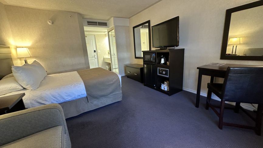 Hotel room with bed, TV cabinet, desk, mirrors and open bathroom doorway.