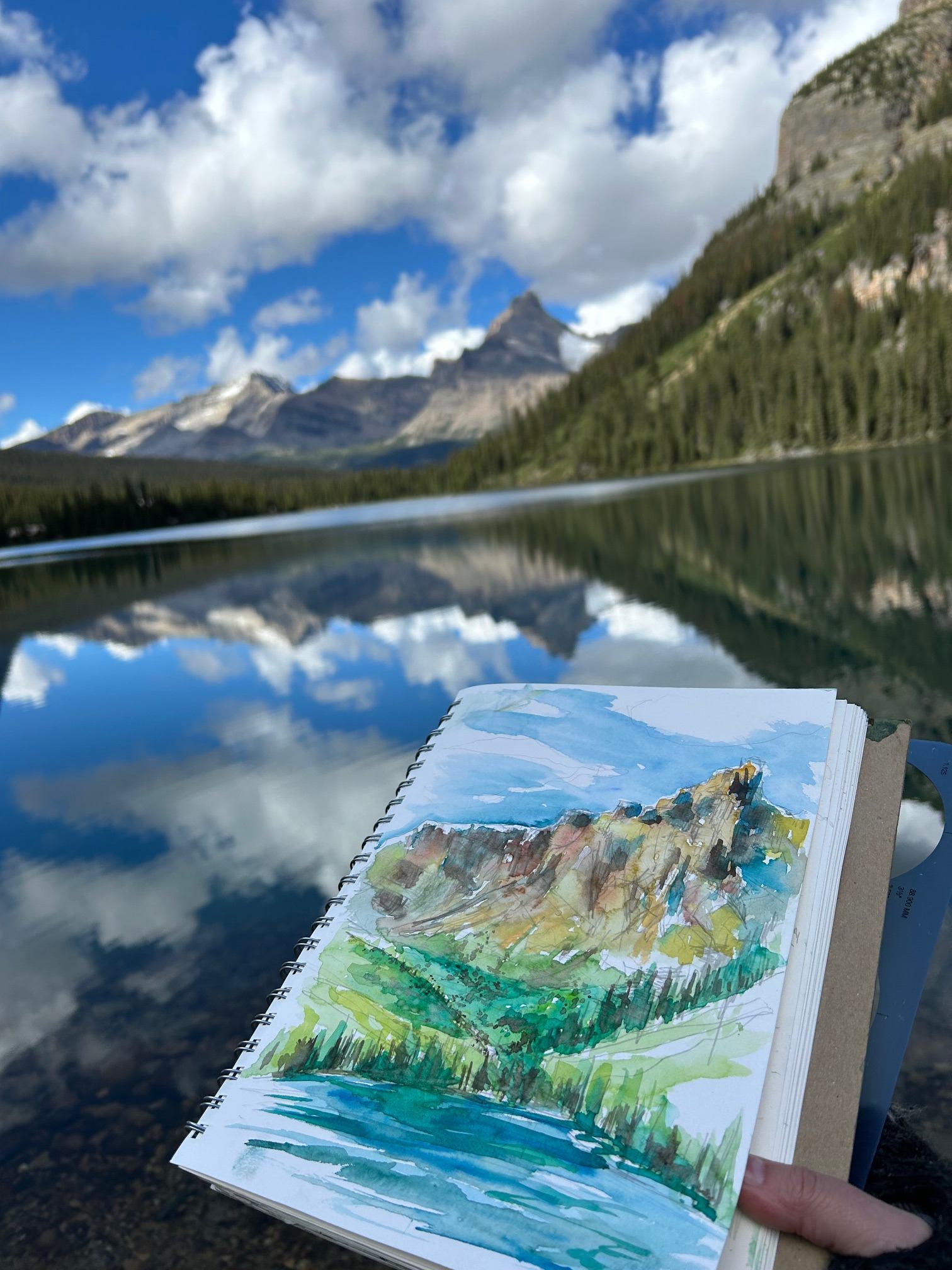 Open sketchbook shows watercolour mountain landscape beside reflective alpine lake and peaks.