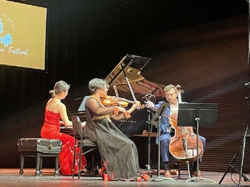 Three musicians perform piano, violin and cello on a concert stage with festival signage.