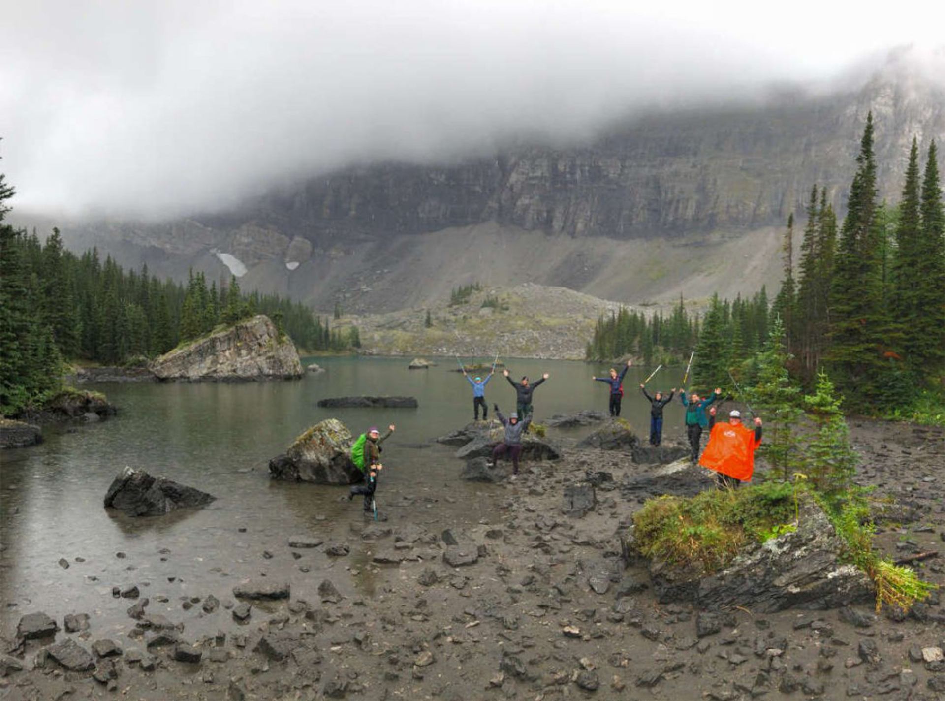 Hikers stand on lakeshore rocks with raised arms, surrounded by evergreens and misty mountains.