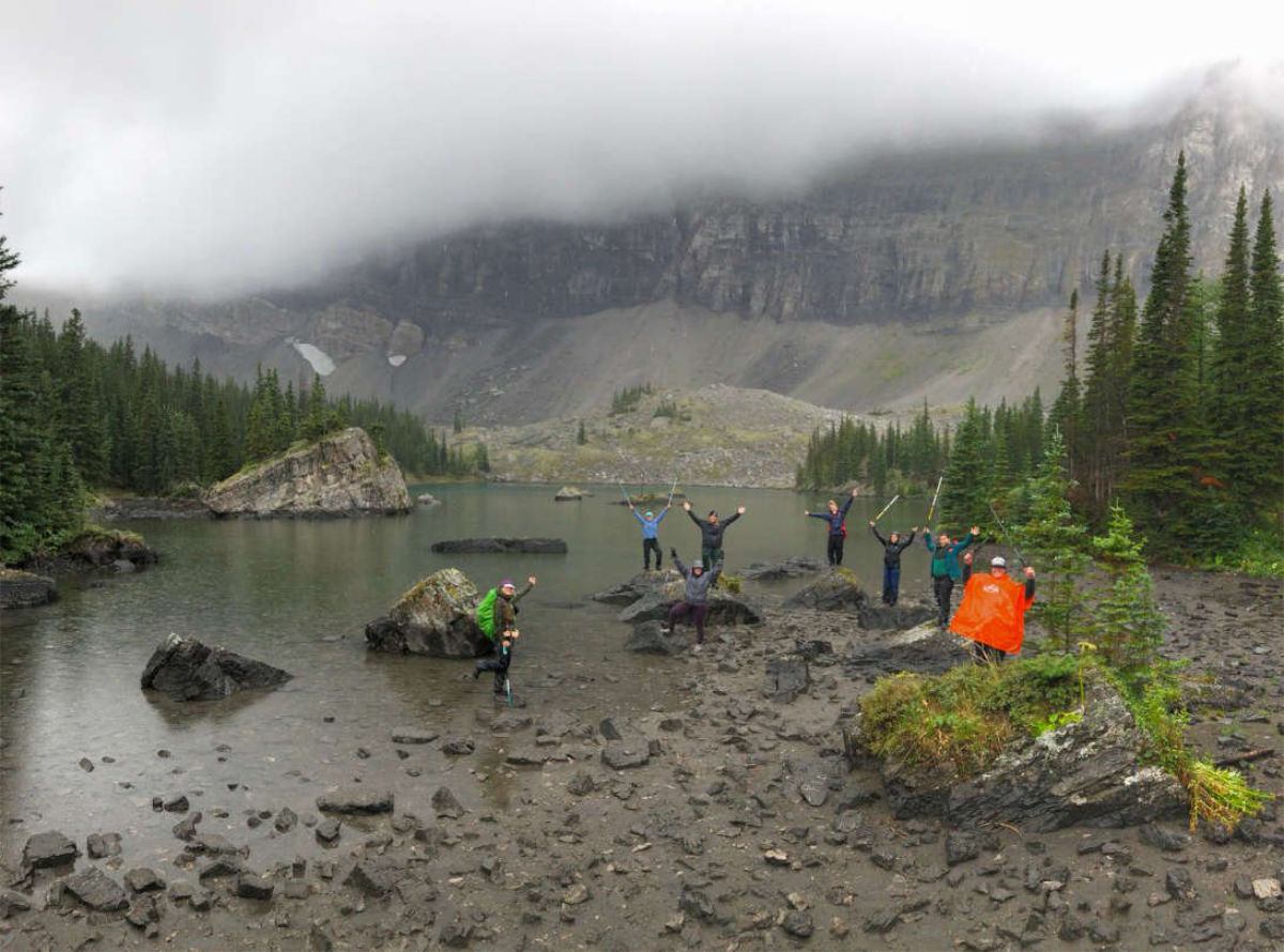 Hikers stand on lakeshore rocks with raised arms, surrounded by evergreens and misty mountains.