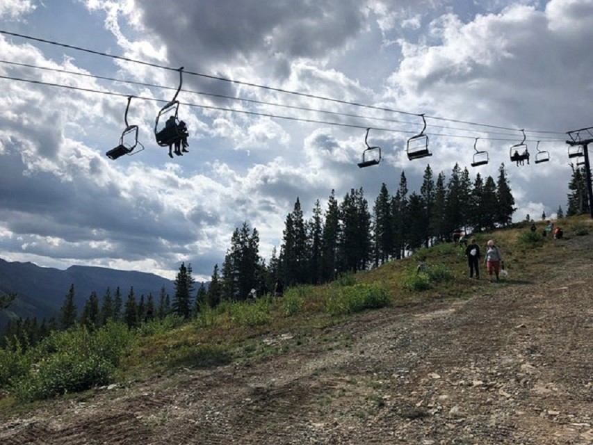 Chairlift passes over a rocky hillside with hikers and dense evergreen forest below.