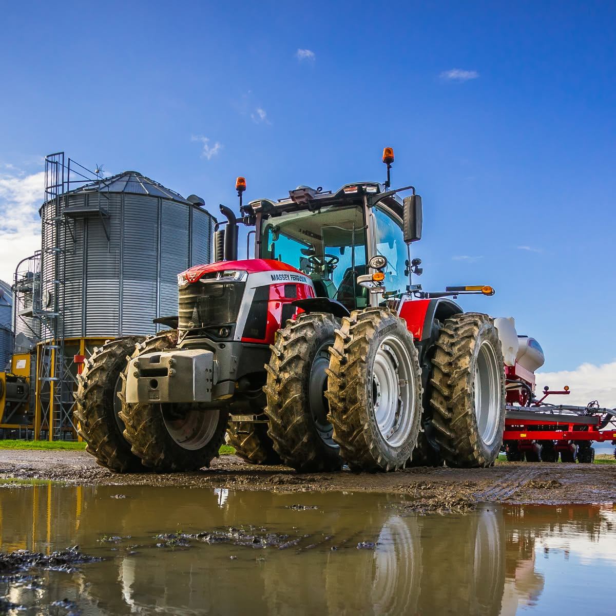 Red Massey Ferguson tractor with dual tyres near metal grain silos and muddy puddle.