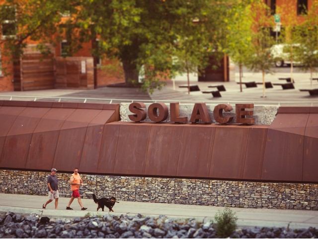 People walk a dog past SOLACE sign on curved metal wall.