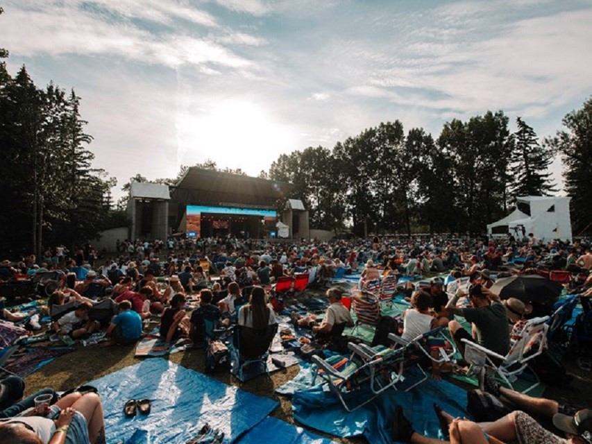A large outdoor audience sits on the grass facing a main stage at the Calgary Folk Music Festival.