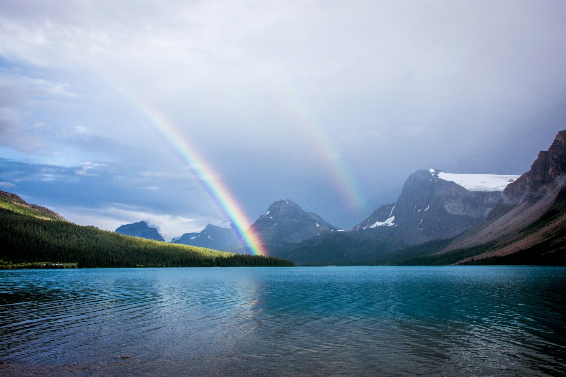 Double rainbow arching over a blue lake with mountains and forest beneath a cloudy sky.