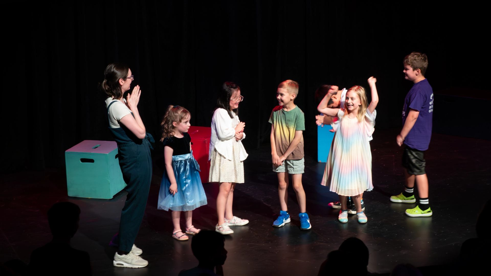 Children stand on a theatre stage with an instructor, performing a group activity.