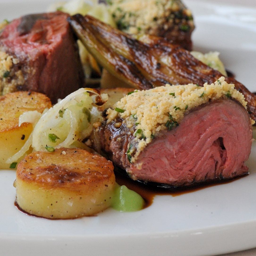 Tender beef fillet with herb crust, served with roasted potatoes, caramelized onions, and a rich sauce. The dish has a gourmet presentation.
