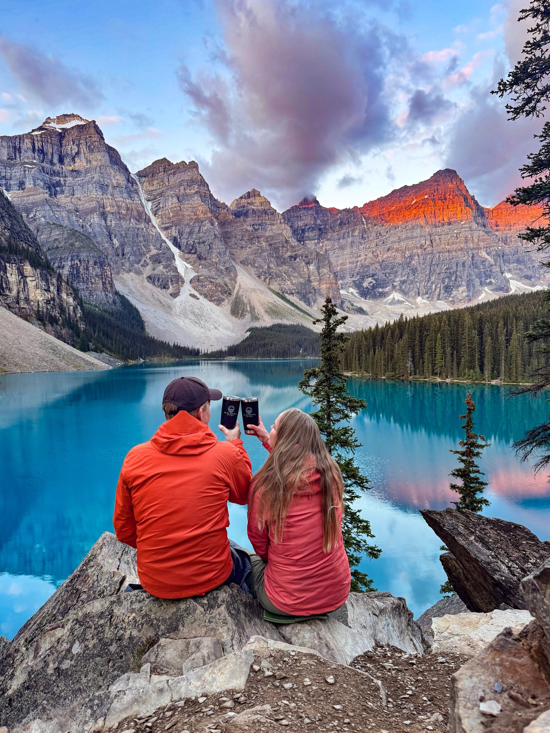 Couple sitting by Moraine Lake watching sunrise-lit mountains.