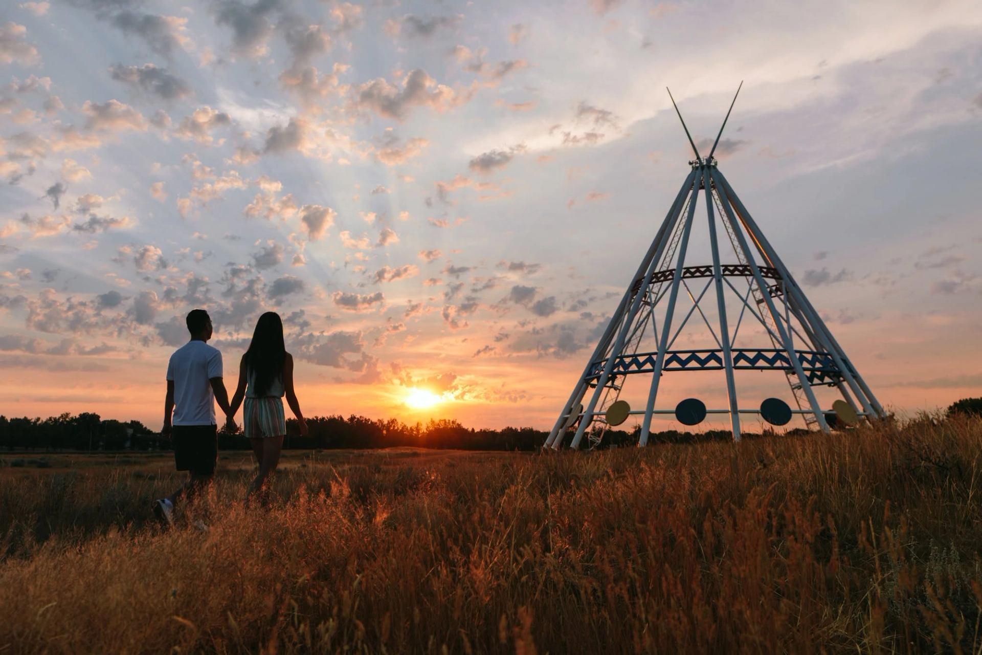 Two people hold hands near a teepee-shaped structure as the sun sets.